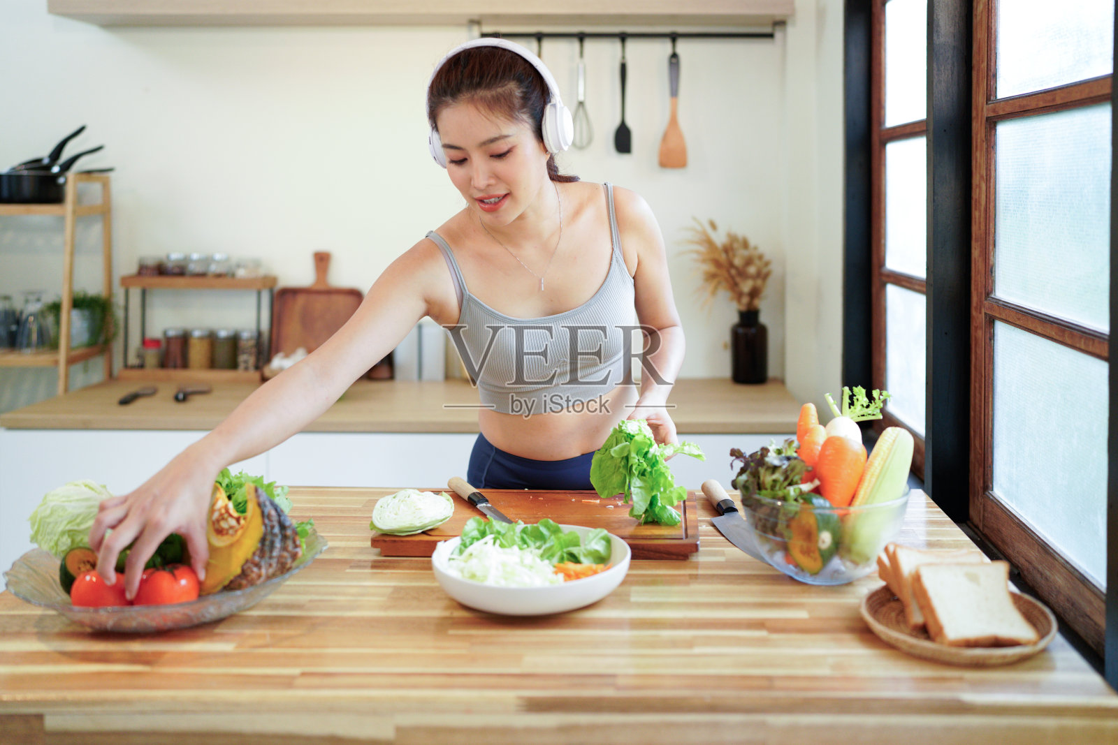 快乐的亚洲健身女性戴着耳机听音乐，正在厨房准备蔬菜沙拉，享受健康的生活方式照片摄影图片