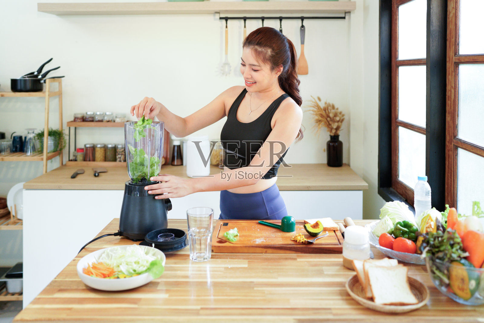 健康食品。一位穿着休闲运动服的女士在家里的厨房里，用搅拌机准备健康的冰沙，并将新鲜的沙拉叶放入搅拌机中制作绿色冰沙。照片摄影图片