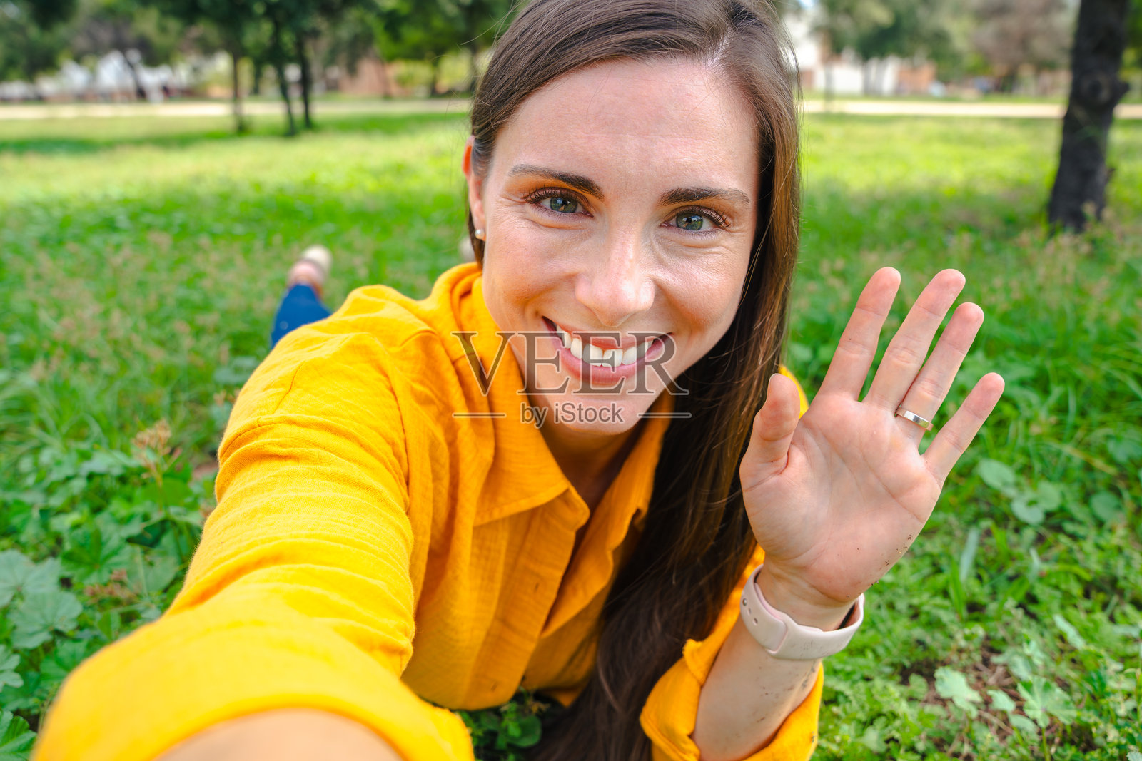 穿着黄色衬衫的女士在户外微笑地自拍，挥着手，背景是绿色公园。照片摄影图片
