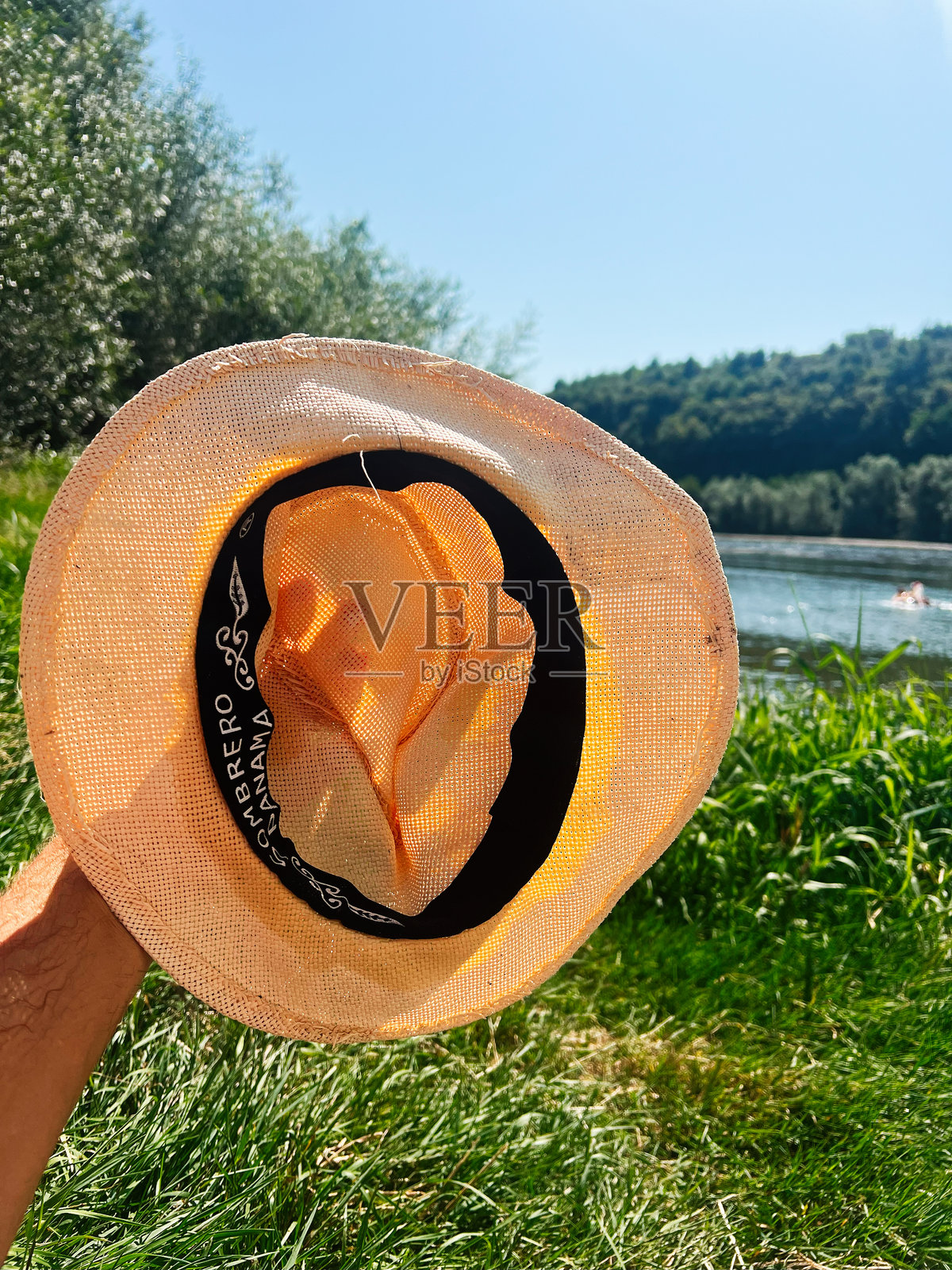 阳光明媚的湖边，大自然环绕中，一只草编巴拿马草帽被户外持有。照片摄影图片