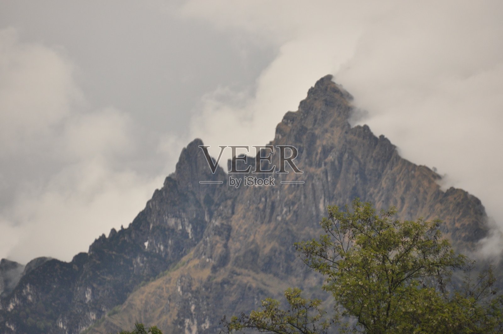 清晨的阳光下，郁郁葱葱的森林旁，雄伟的山峰笼罩在云层之中，天空布满乌云。照片摄影图片
