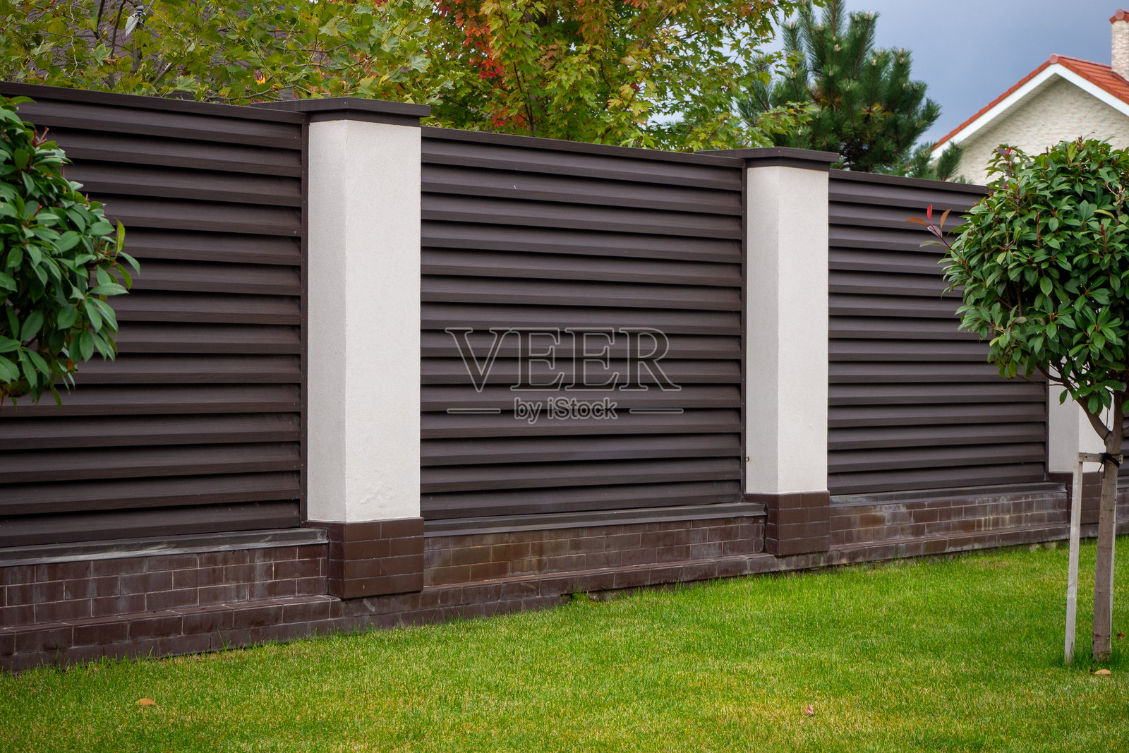 现代深棕色复合材料围栏，配有白色柱子和砖砌基座，环绕着绿色草坪。用于说明隐私屏风、物业边界和现代住宅入口设计。真实照片照片摄影图片