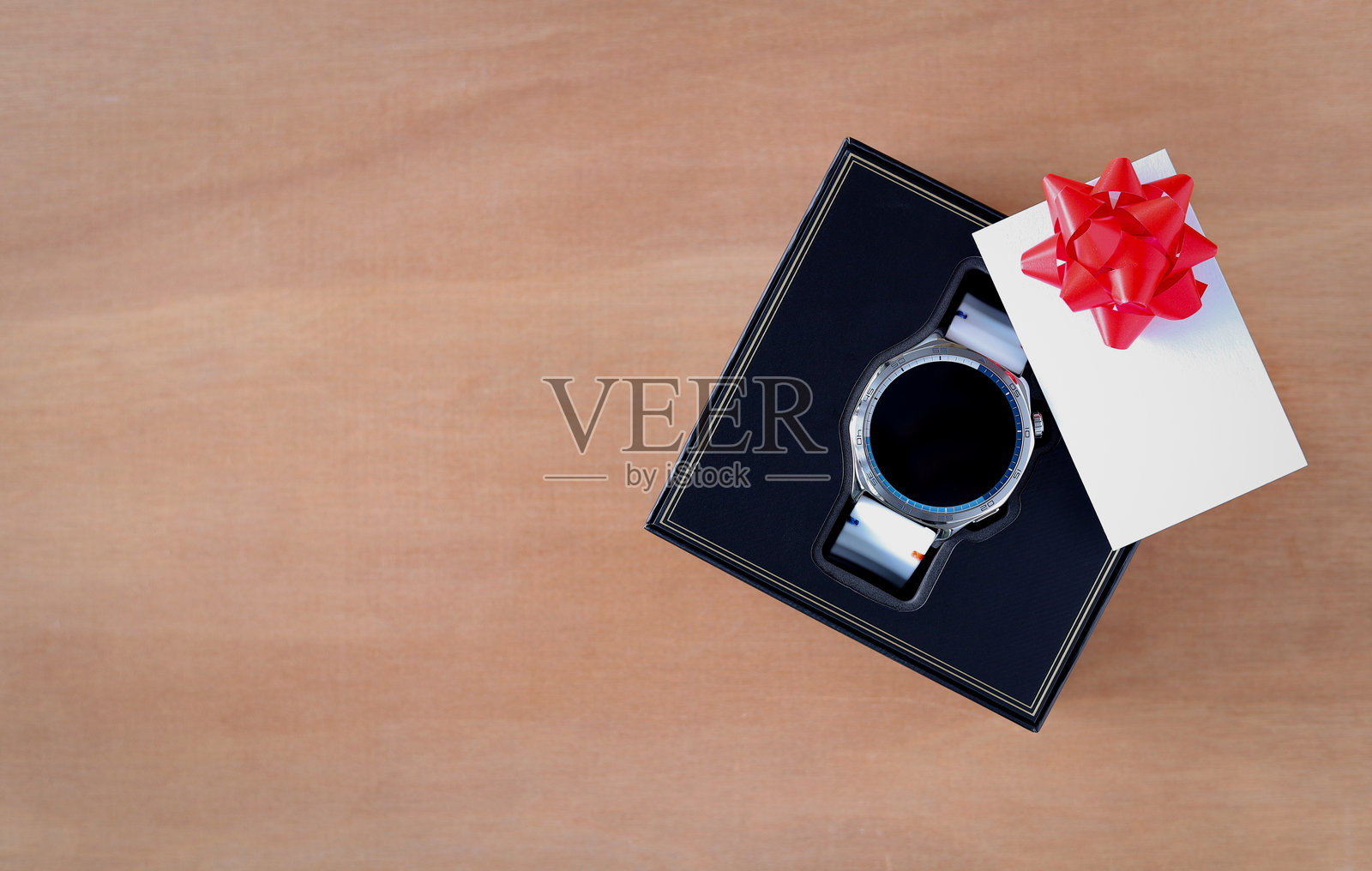 俯视角度拍摄的现代智能手表，置于一个奢华的黑色礼品盒中。盒盖上放着一张带有红色蝴蝶结的空白卡片，旁边留有供书写祝福语的空间，背景为木质桌面。非常适合节日或庆祝活动。照片摄影图片