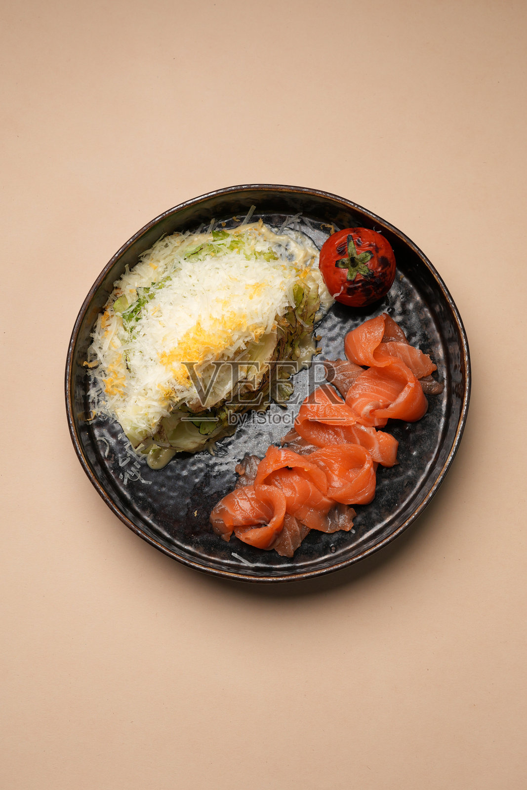 深色乡村风的盘子里盛着烟熏三文鱼片，旁边配有撒了奶酪的沙拉块和烤番茄，背景为米色。照片摄影图片