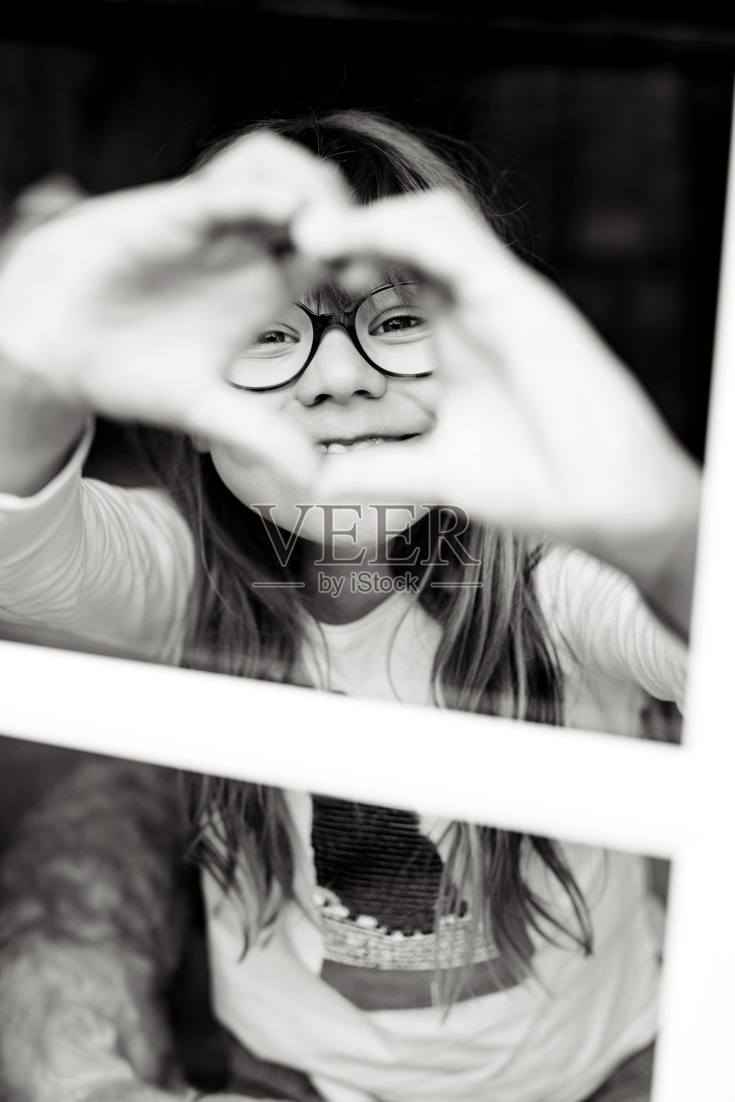 戴眼镜的可爱小女孩，双手比心，透过窗户看向外面，在温馨的家中表达爱与喜悦照片摄影图片