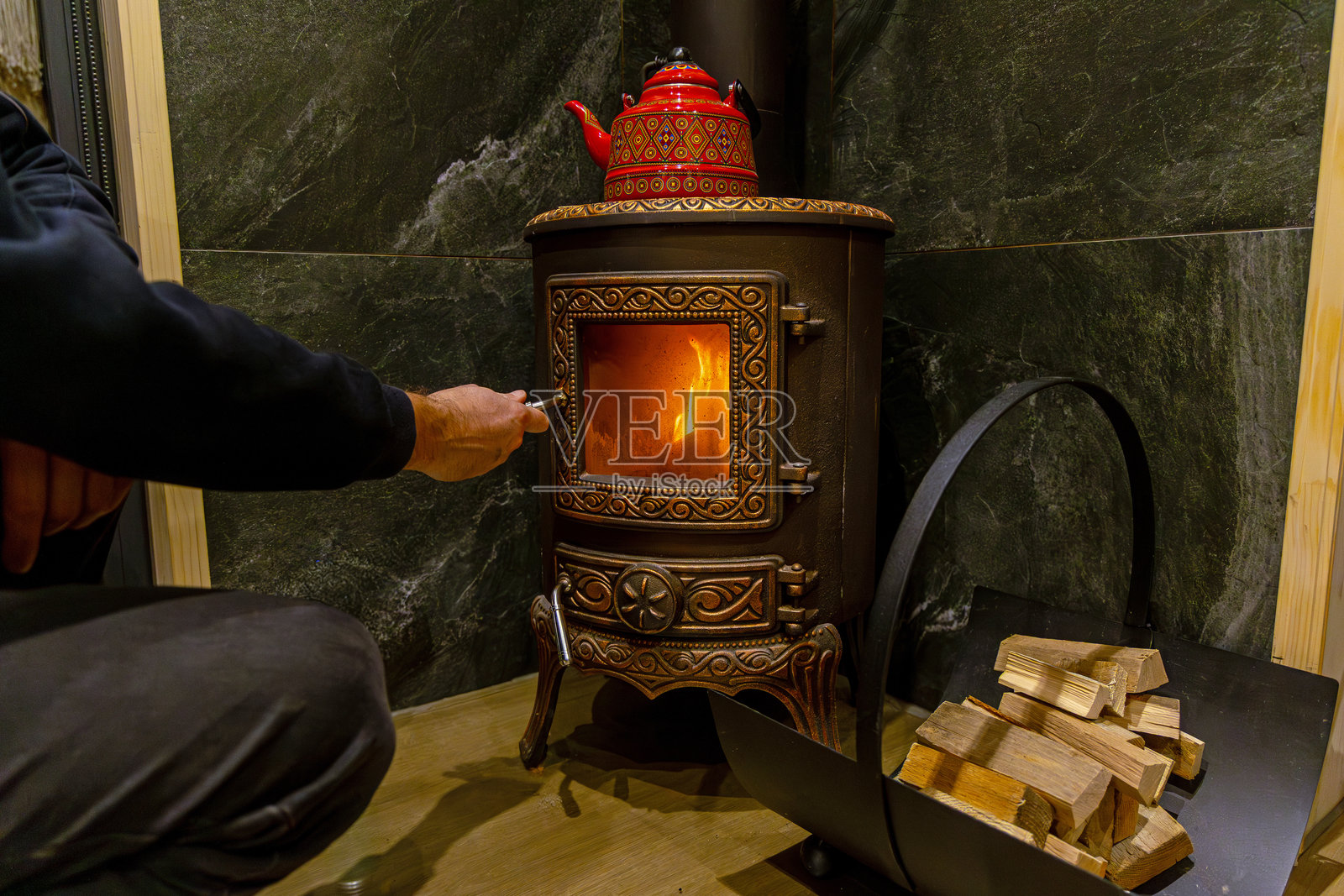 男人点燃装饰性的铸铁炉，炉内有木柴，炉顶放着红色水壶，旁边堆放着木柴，室内场景。照片摄影图片
