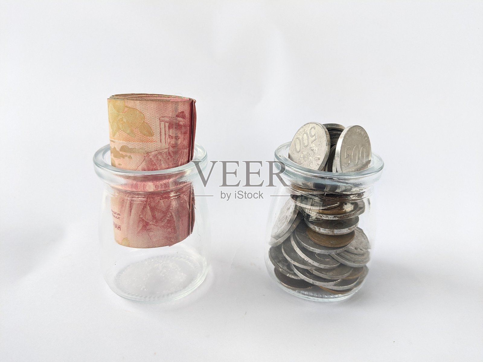 照片展示了两个放在白色背景上的小型透明玻璃容器。左边的容器里装着一张整齐卷起的纸币。照片摄影图片