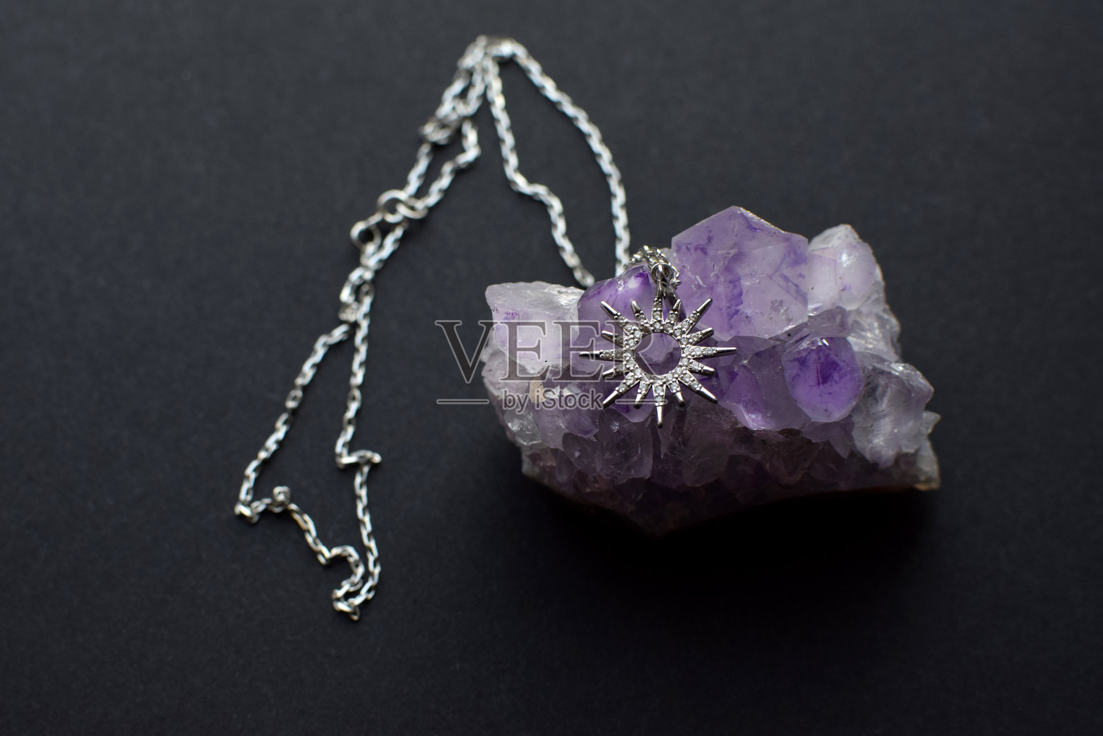 深色背景上，镶嵌宝石的太阳或星星形状的银质吊坠挂在链子上，吊坠位于紫水晶簇之上。照片摄影图片