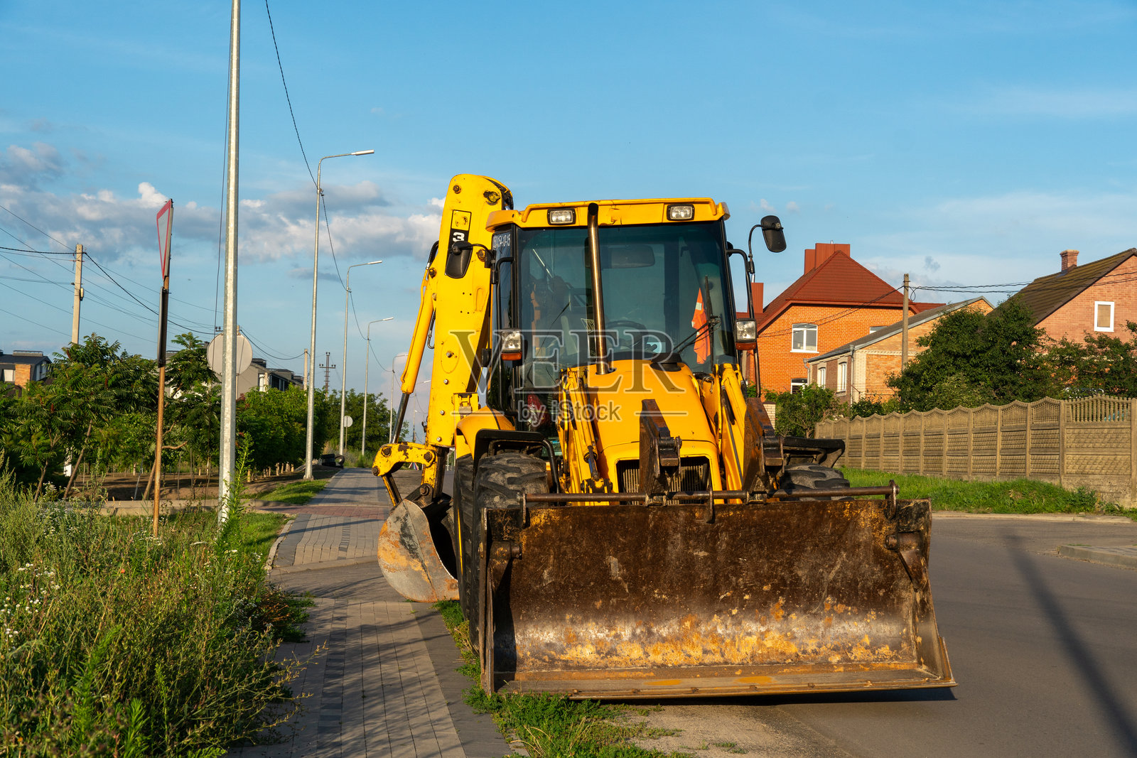 一辆大型黄色挖掘装载机停在郊区街道上，靠近住宅房屋，天空湛蓝晴朗。照片摄影图片