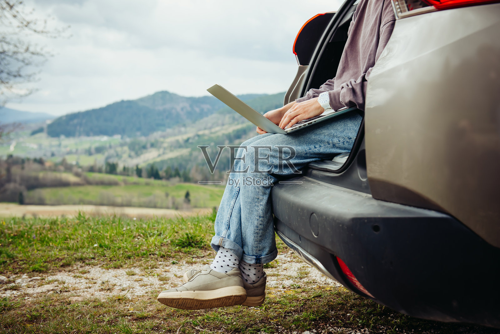 一位选择慢节奏生活的女孩在旅途中，坐在汽车后备箱里用笔记本电脑工作，体验着工作与旅行相结合的理念。照片摄影图片