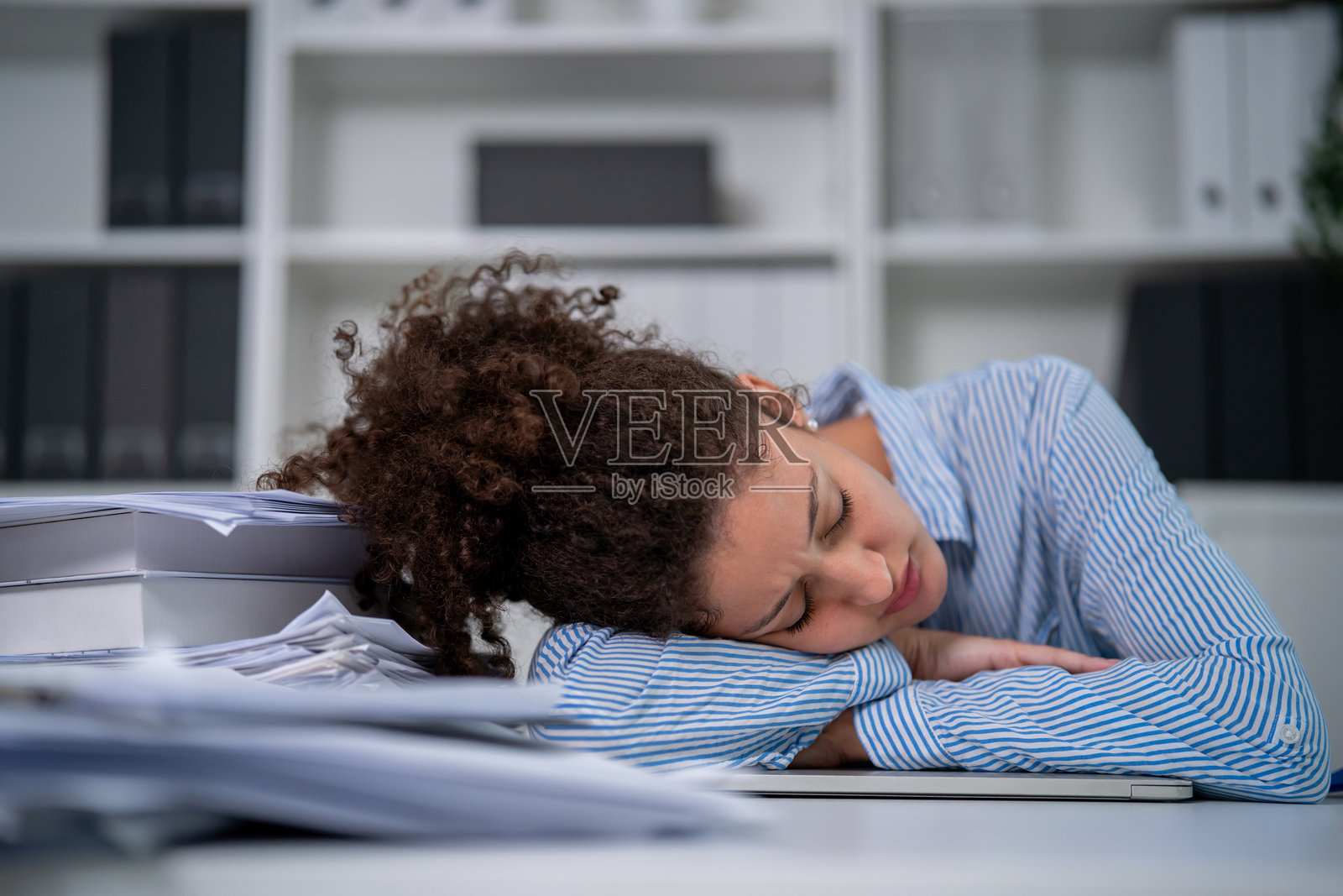 疲惫的年轻拉丁裔女商人正在办公室加班。一位迷人的忙碌女自由职业者感到疲倦和疲惫，将头靠在桌子上，在舒适的工作场所睡着了。照片摄影图片