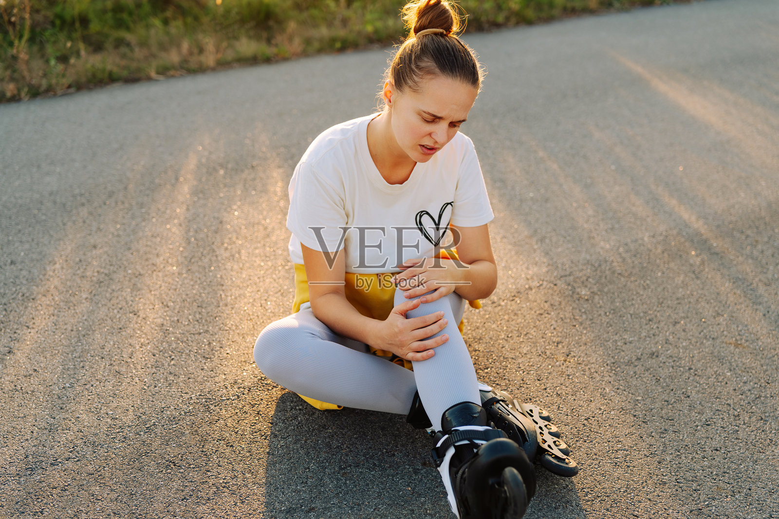 身穿运动服的女子痛苦地跪坐在地上，抱着膝盖，夕阳将长长的影子投射在空旷的道路上。照片摄影图片