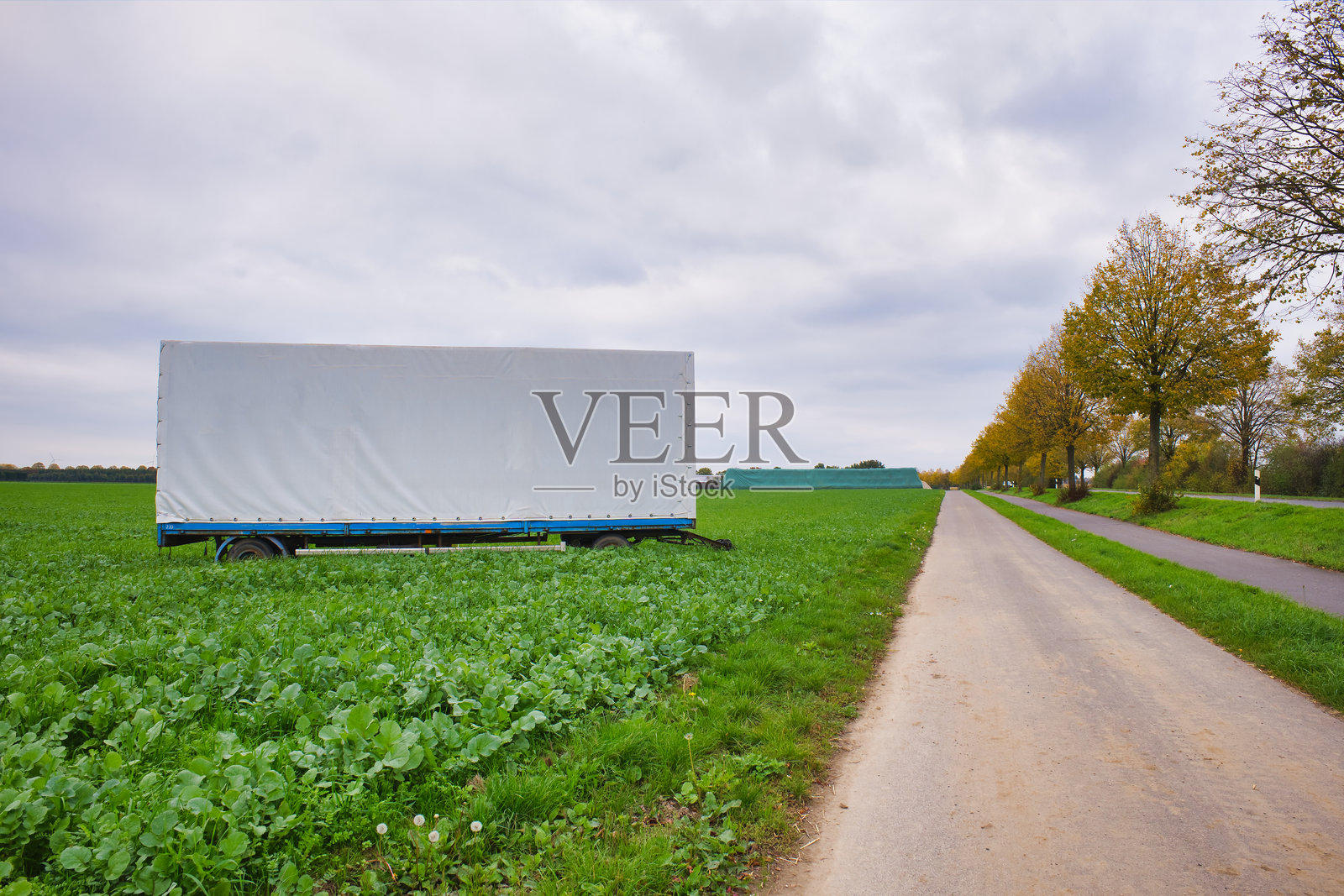 白色的拖车停在乡村公路边，周围是绿色的田野，远方地平线上排列着金色的秋季树木，天空布满云彩。照片摄影图片