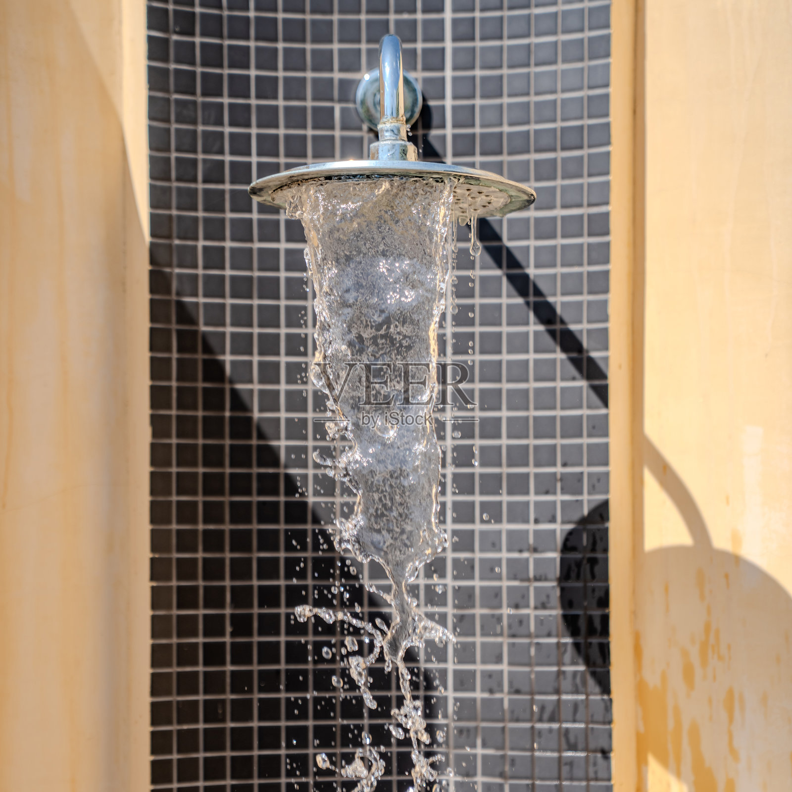 水从圆形的金属淋浴喷头流出，背景是瓷砖墙壁，阳光突出了水的纹理和户外的环境。照片摄影图片