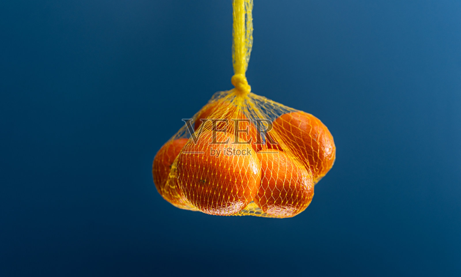 鲜艳的橘子悬挂在黄色的网兜中，背景为深蓝色。照片摄影图片
