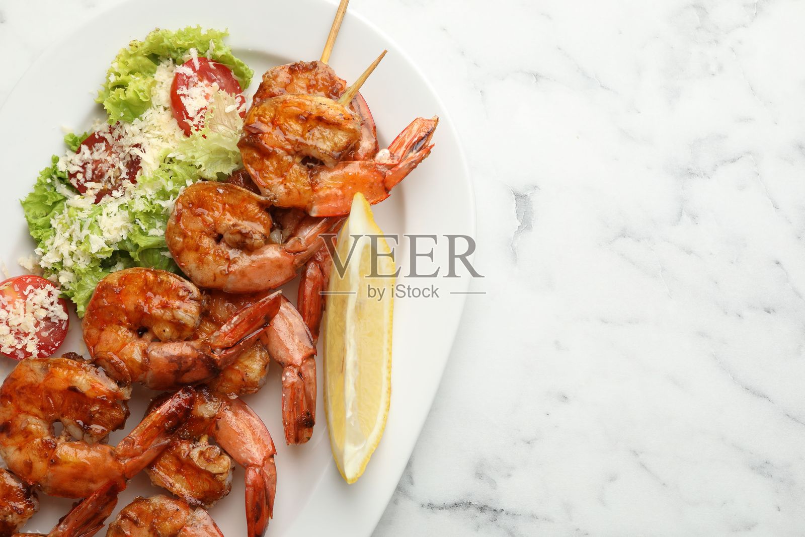 烤虾串、沙拉和柠檬片放在白色大理石桌上，俯视视角，留有文字空间。美味的海鲜菜肴照片摄影图片
