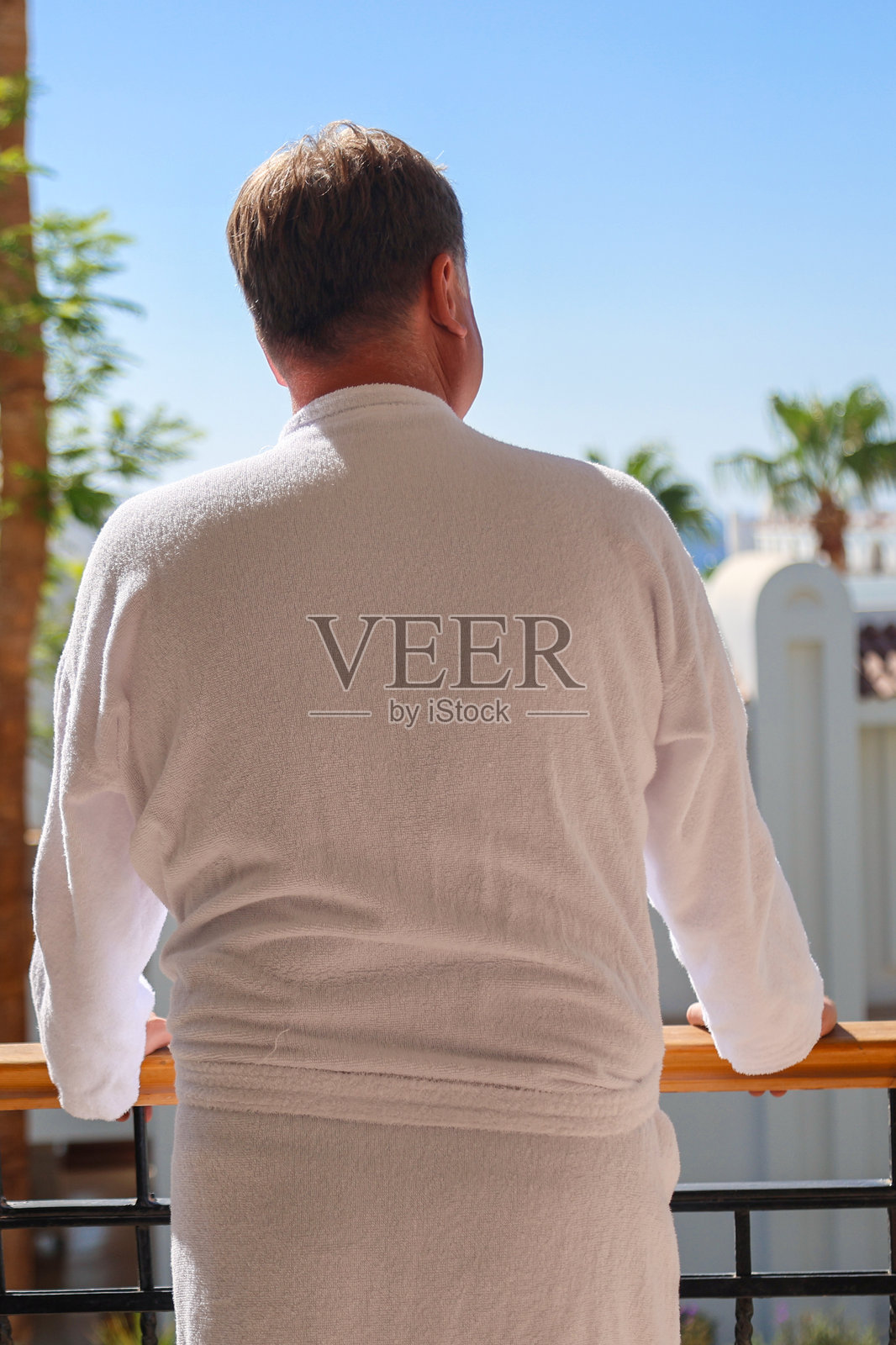 身着浴袍的男子背身站立在豪华住宅的露台上，眺望着大海和棕榈树照片摄影图片