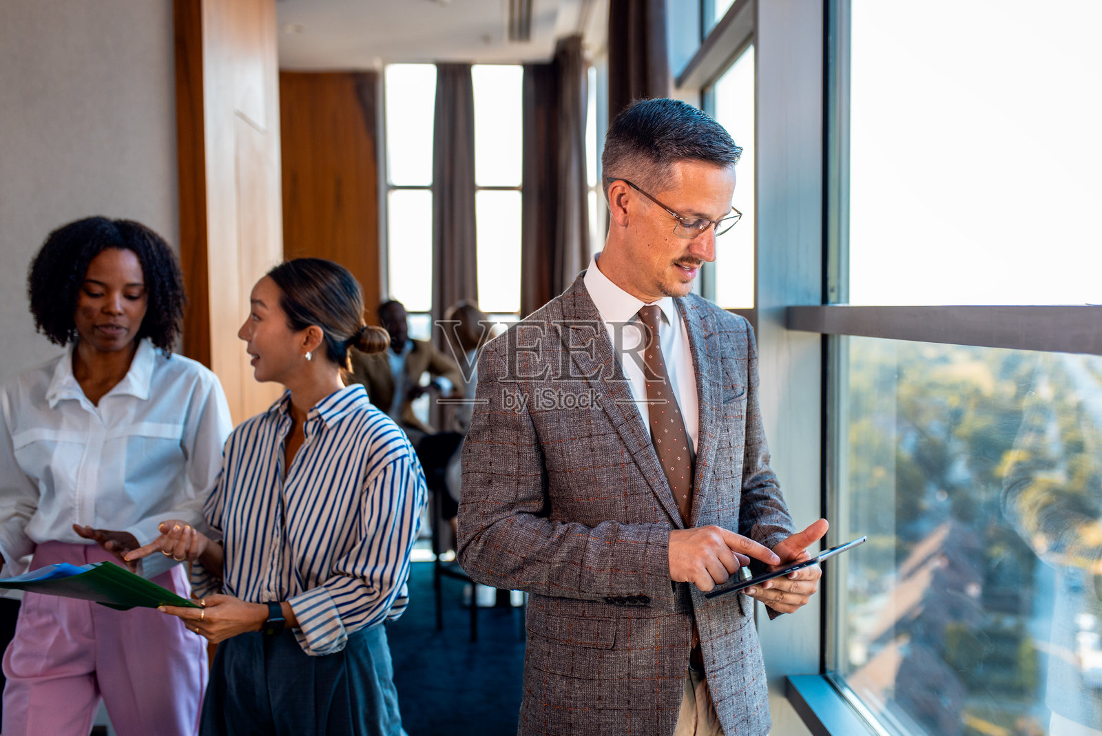 商务人士站在办公室靠窗的位置，使用平板电脑。照片摄影图片