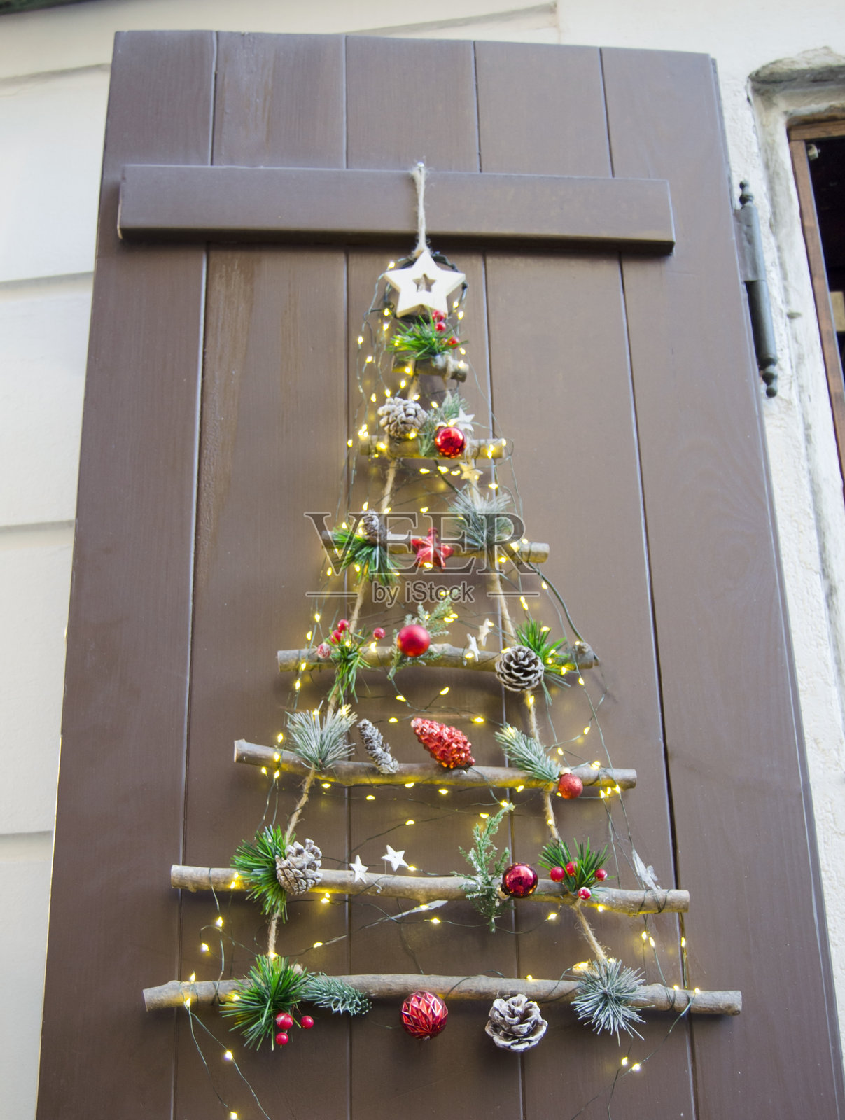 用木枝、松果和节日红色装饰品手工制作的创意圣诞树，用温暖的仙女灯装饰，挂在质朴的木门上。照片摄影图片