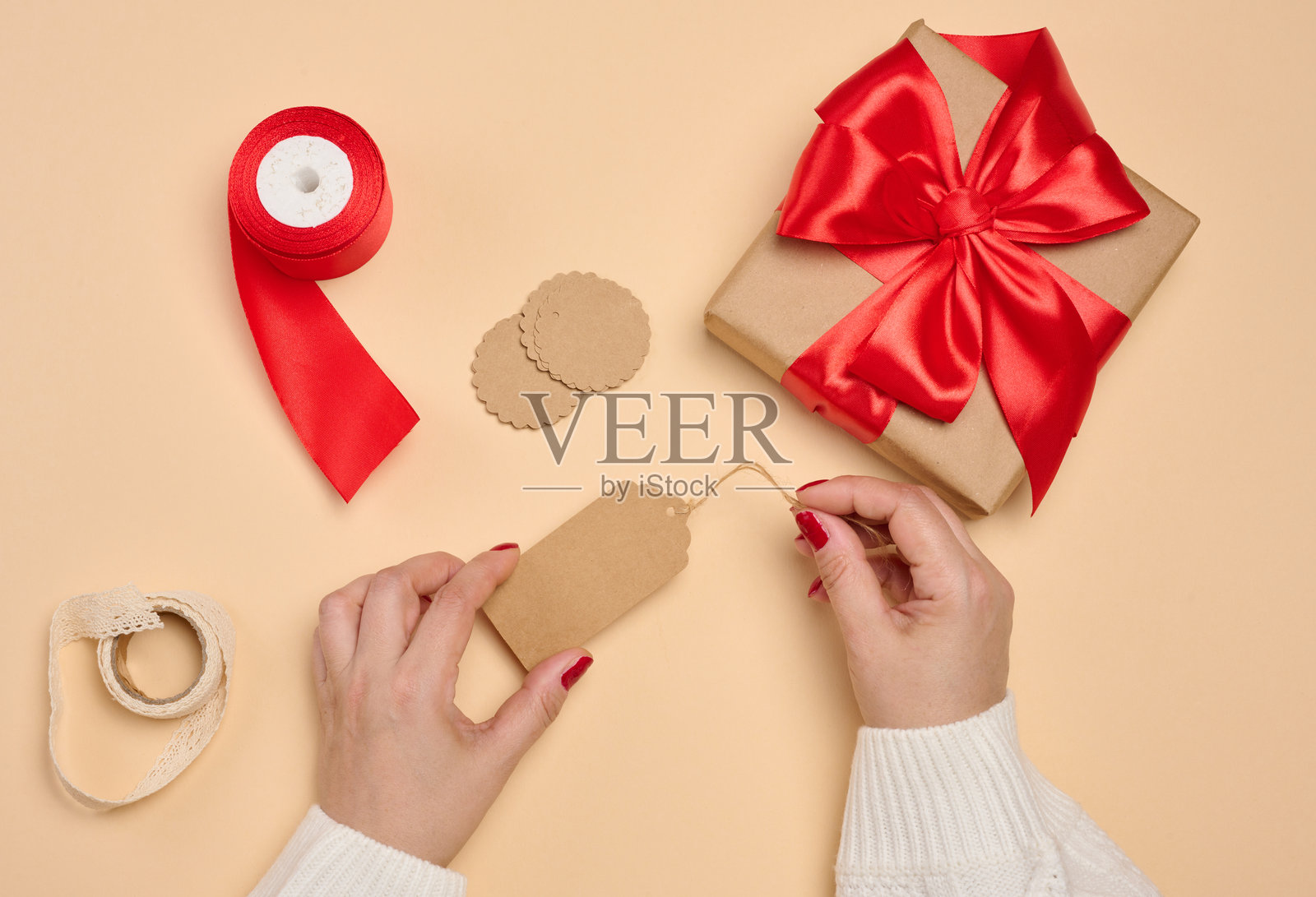 穿着白色毛衣的女士双手拿着一个礼物盒，礼物盒用牛皮纸包装，上面系着一个红色缎带蝴蝶结，还有吊牌和一卷丝带。照片摄影图片