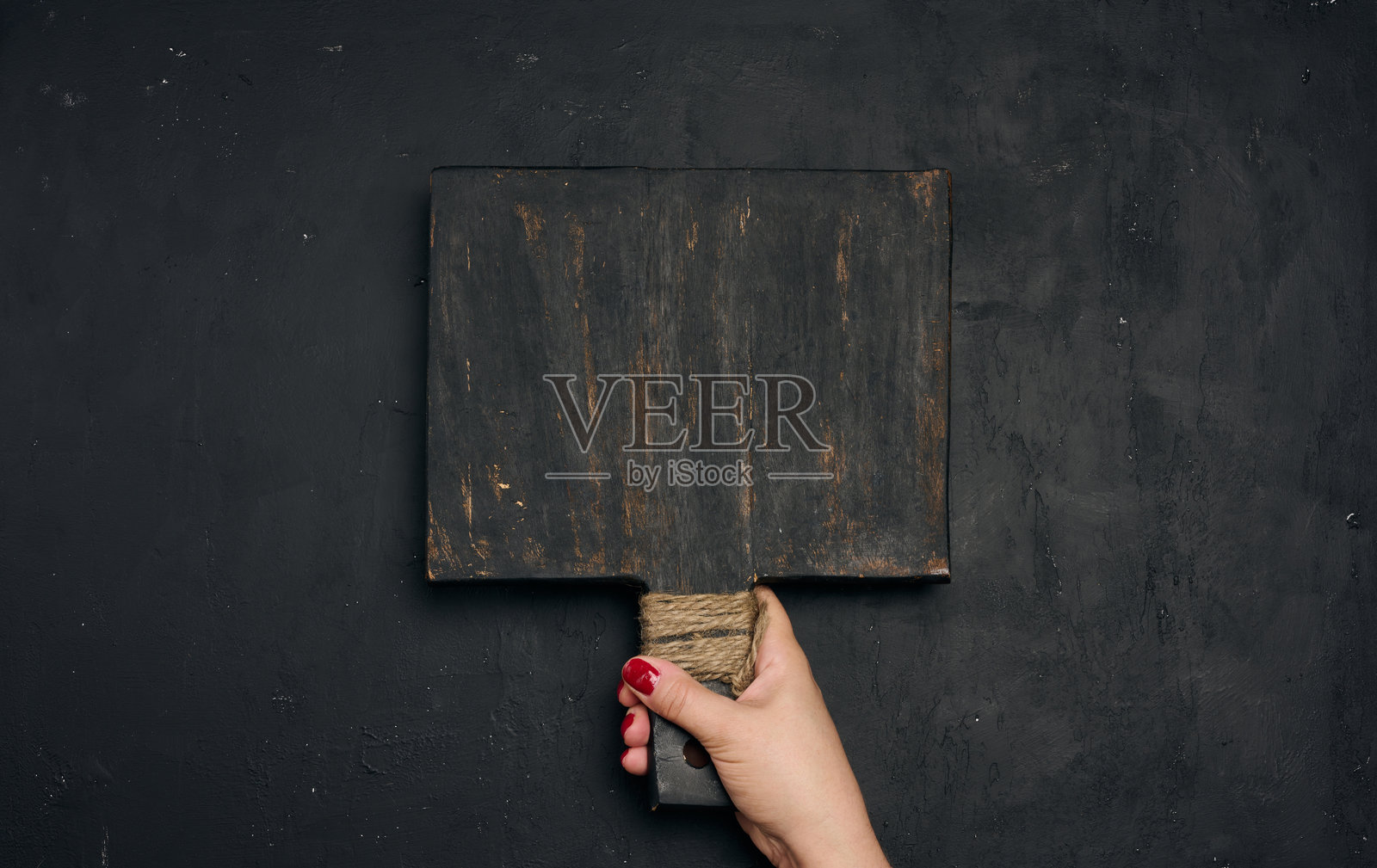 一只女人的手握着一个旧的、黑色的木制厨房砧板的把手，砧板用麻绳缠绕着，背景是黑色的纹理。照片摄影图片