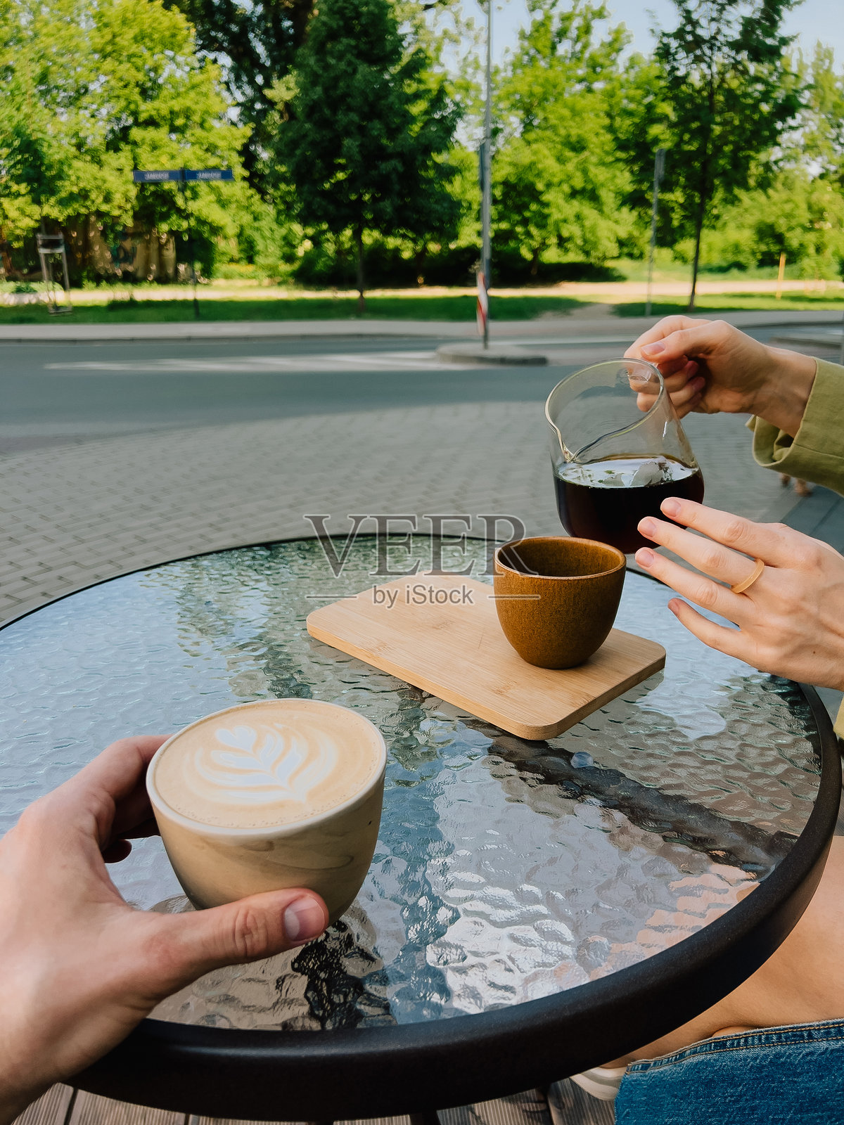 第一人称视角：手持咖啡，在户外咖啡馆的玻璃桌上倒茶，背景是阳光明媚夏日里充满活力的城市公园，营造出完美的放松时刻。卡布奇诺。照片摄影图片