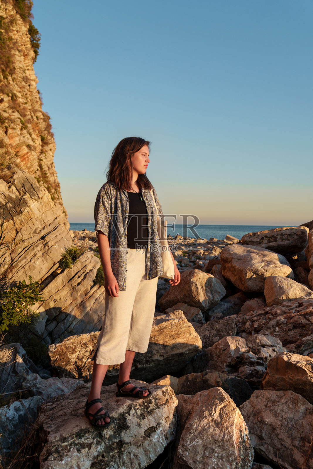 日落时分，一位女孩站在高崖斜坡附近的岩石上海边，眺望海景和山景的全景照片摄影图片