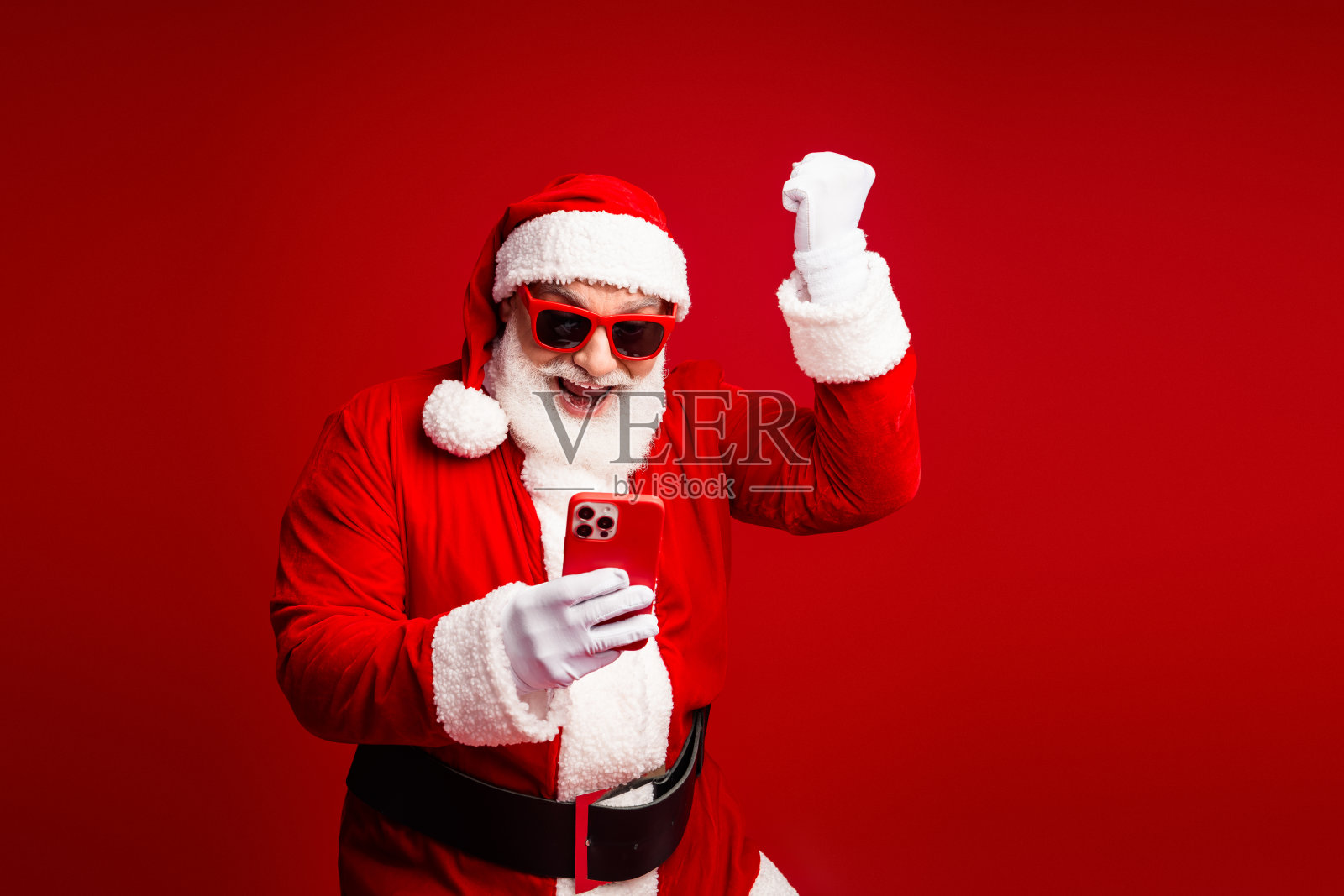 圣诞老人在红色的工作室里，戴着圣诞帽和太阳镜，在平安夜庆祝活动中用智能手机欢快地自拍。照片摄影图片