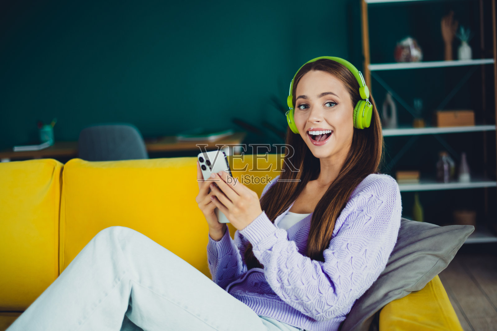 年轻女子戴着绿色耳机，在家中明亮的黄色沙发上放松，对着手机微笑，场景是现代舒适的客厅照片摄影图片