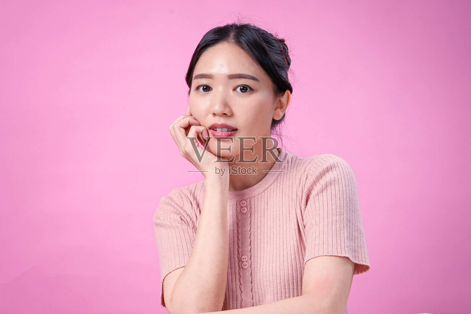 一位留着整洁发髻黑发的年轻亚洲女性，身穿罗纹粉色短袖上衣，在粉色纯色背景前，手托着下巴，面带灿烂的笑容，姿势愉快照片摄影图片