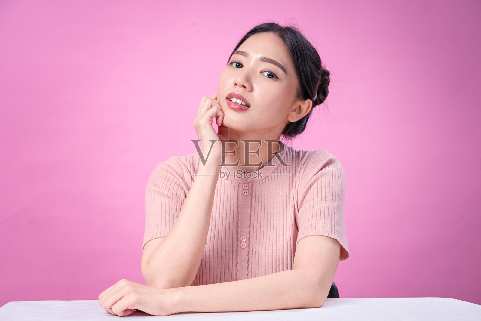 一位年轻的亚洲女性，留着整齐的黑色盘发，身穿罗纹粉色短袖上衣，面带灿烂的微笑，一只手托着下巴，背景为纯粉色，姿态欢快。照片摄影图片