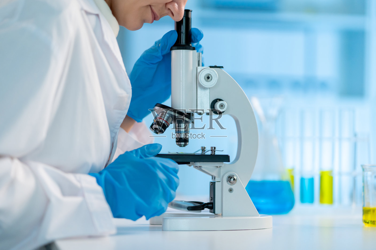 近距离拍摄一位身穿白色实验服的女科学家在实验室工作。一位有吸引力的年轻研究技术员正在使用显微镜进行医学研究，专注于现代科学实验室的样本。照片摄影图片