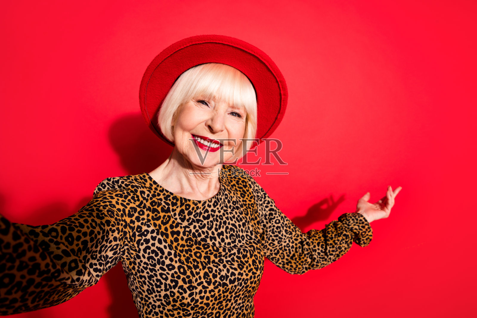 一位快乐、友善、漂亮的年长女士伸出手表示欢迎，邀请您微笑自拍，背景为红色照片摄影图片