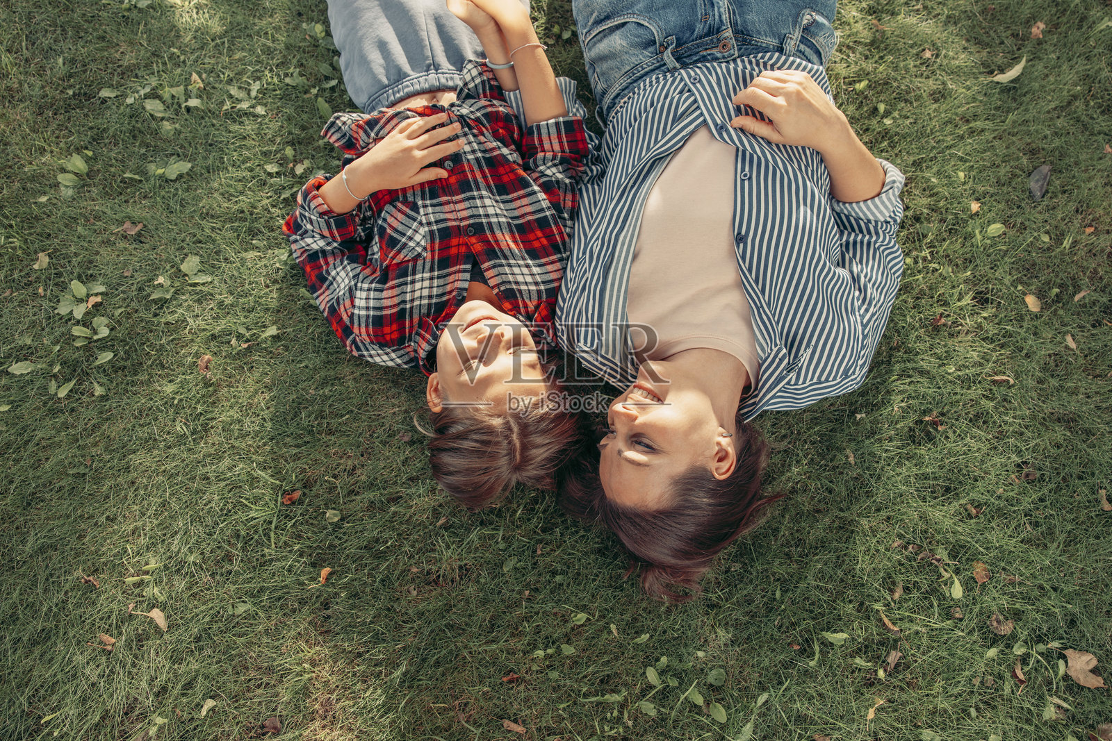 两个女人躺在绿草地上，微笑着享受彼此的陪伴。一个留着长棕发，穿着格子衬衫，另一个留着中长棕发，穿着条纹衬衫。照片摄影图片
