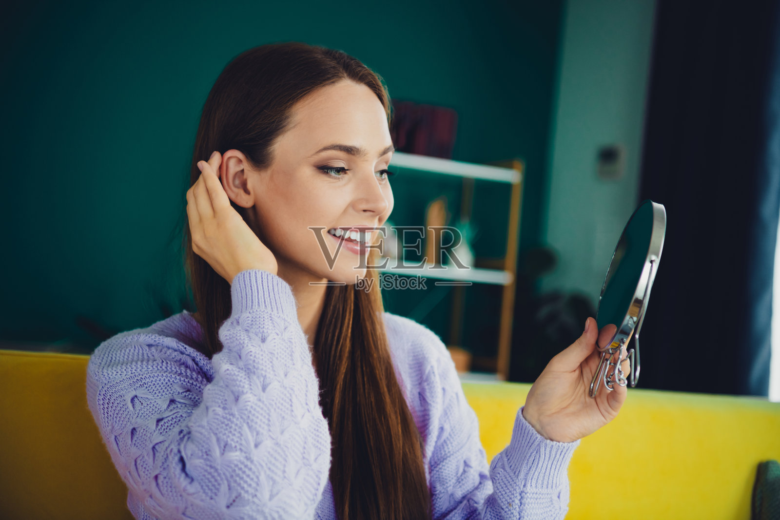 年轻女子穿着薰衣草色毛衣，在家中明亮的客厅里，手持镜子审视自己，展现出休闲的美丽生活方式和温馨的室内氛围照片摄影图片