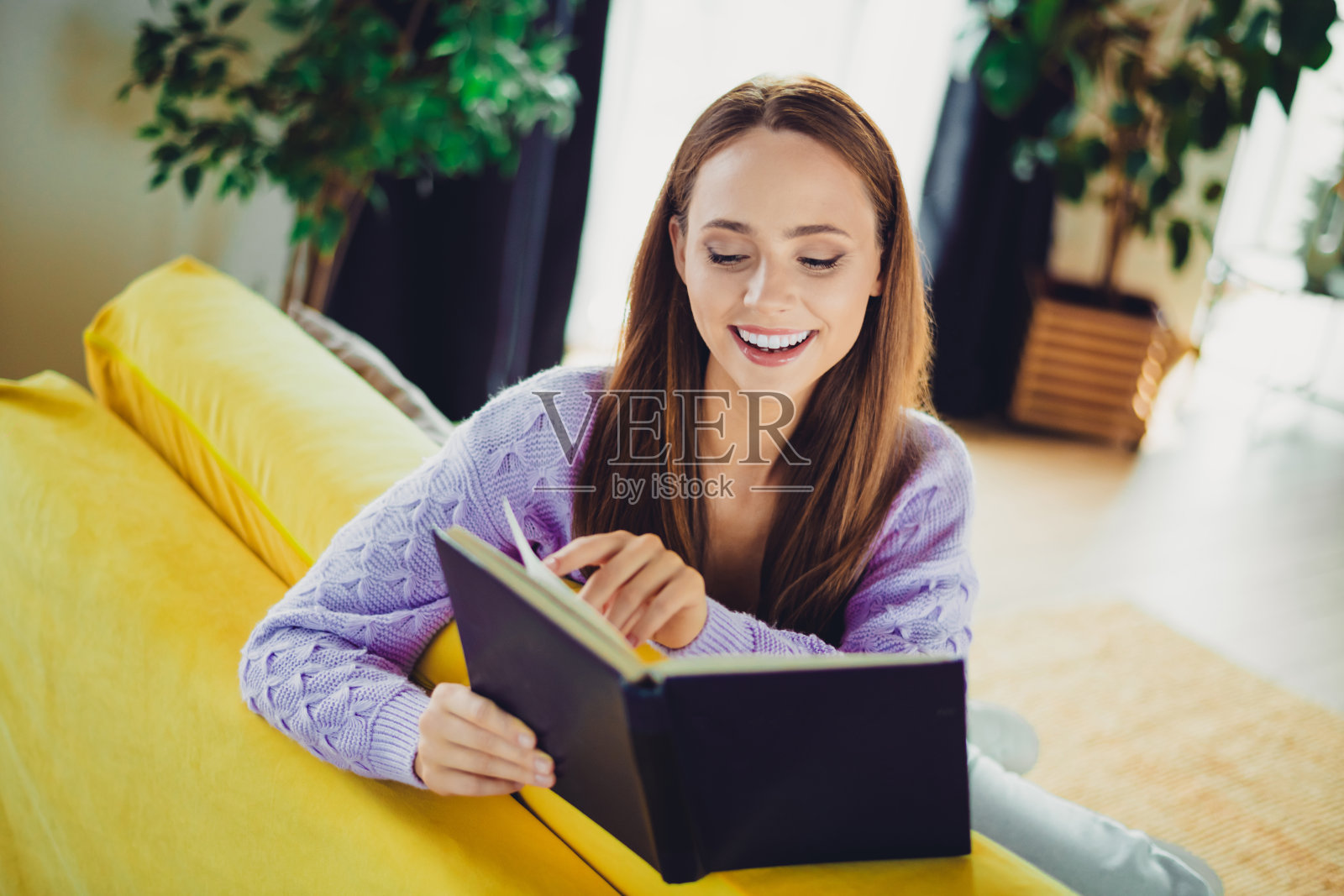 一位快乐的年轻女子在家中舒适的客厅里，放松地坐在亮黄色的沙发上，阅读着笔记本。照片摄影图片