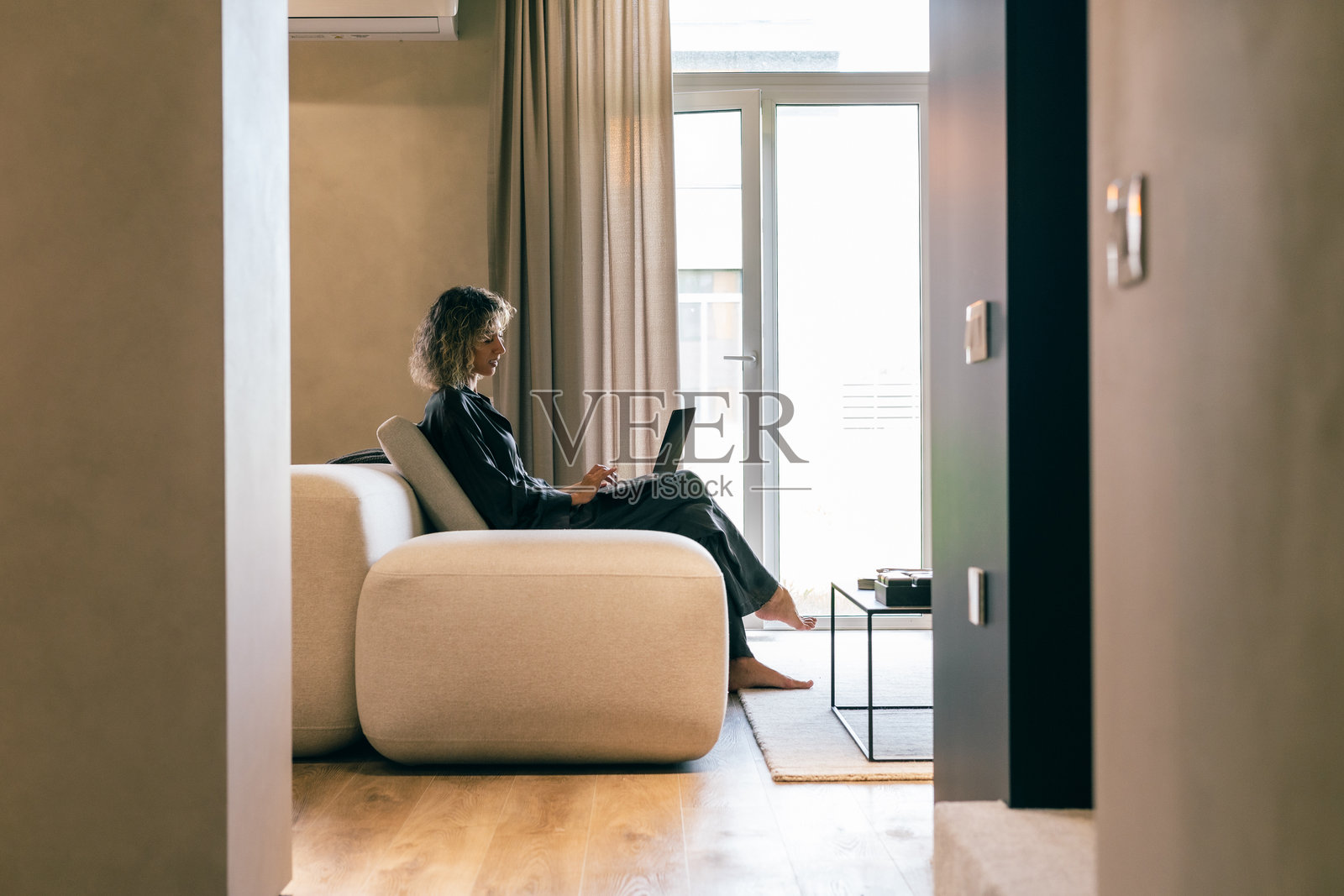 女子舒适地坐在现代沙发上，在自然光下用笔记本电脑在家工作照片摄影图片