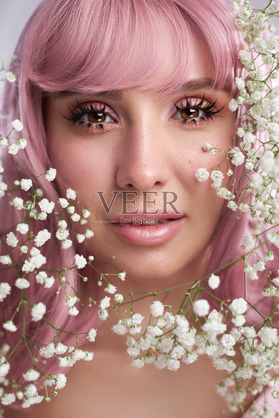 拥有柔和粉色头发的年轻女子温柔地望着镜头，周围环绕着精致的白色花朵，在宁静的氛围中散发出平静与美丽的气息。照片摄影图片