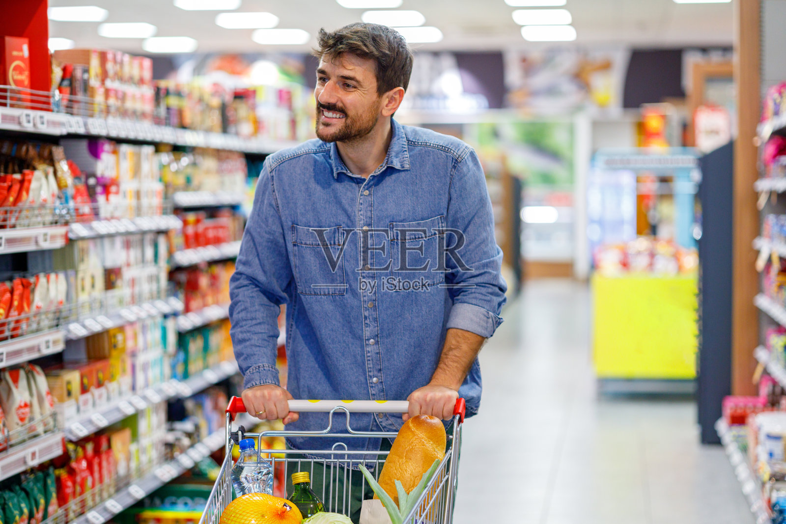 一位快乐的年轻男子正在熙熙攘攘的超市里购物，他推着一辆装满新鲜农产品的购物车。照片摄影图片