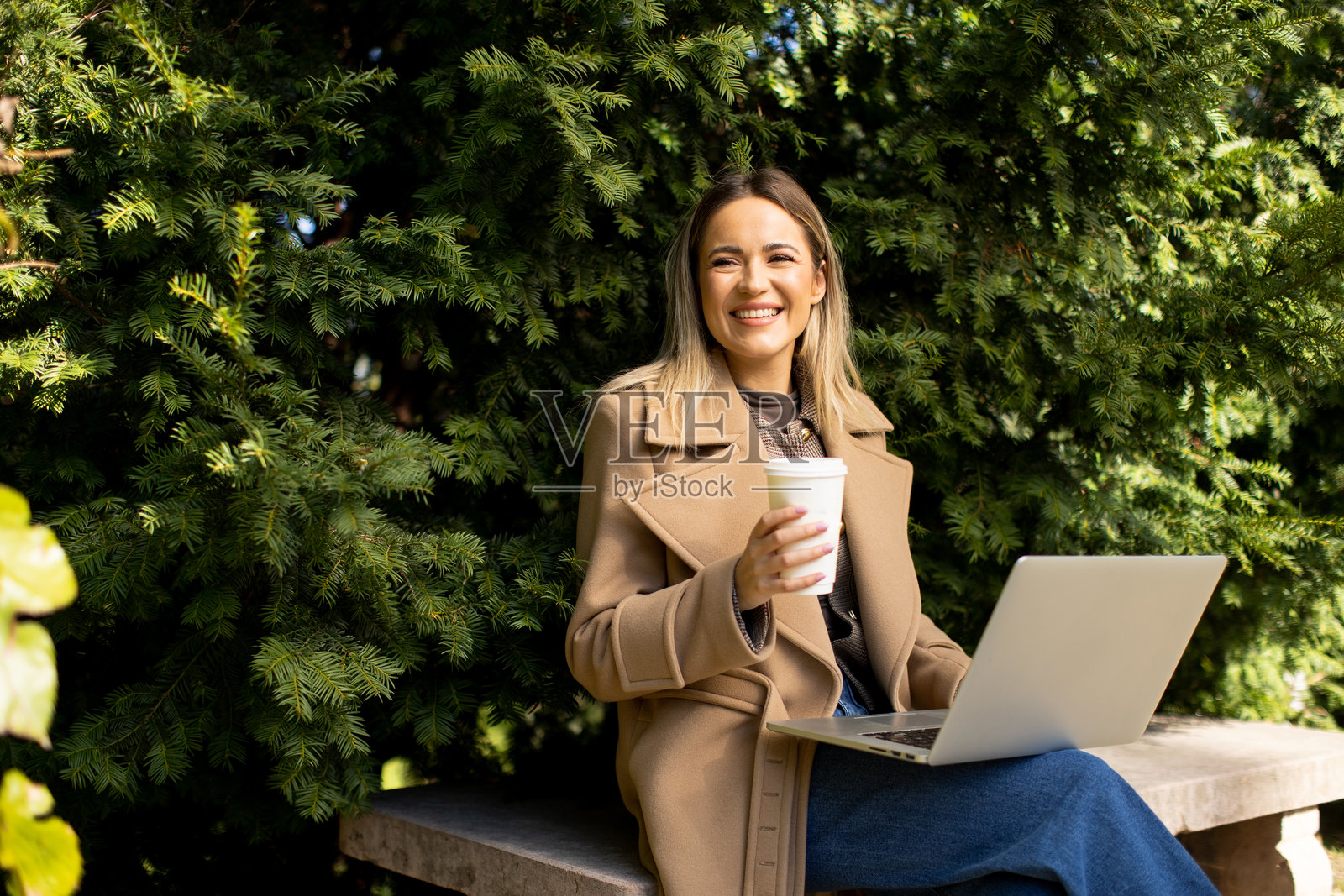 阳光明媚的户外，女子在郁郁葱葱的绿植间用笔记本电脑工作，旁边放着一杯咖啡照片摄影图片