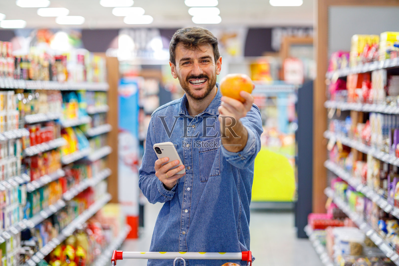 一名男子在超市里，一手拿着手机，一手拿着新鲜水果，微笑着，正在选购食品和健康食品。照片摄影图片