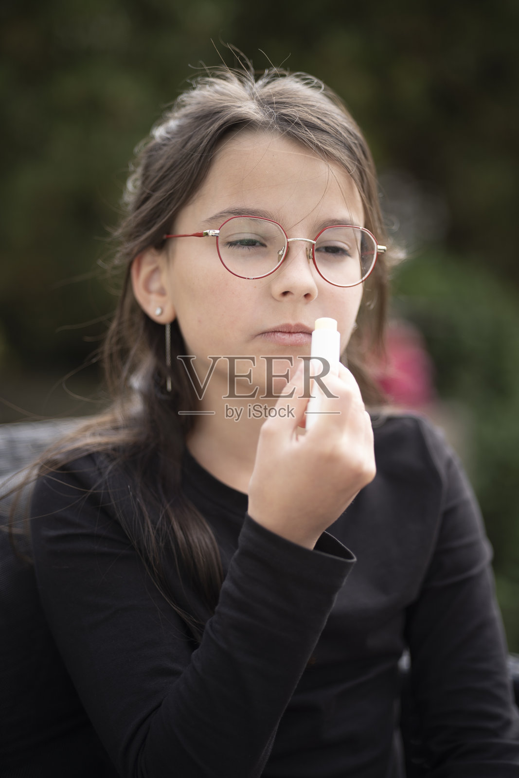 戴眼镜的黑发女孩穿着长袖衣服，坐在户外，在模糊的自然背景下涂抹口红，柔和的日光，可复制空间。象征成长、好奇心和自我表达。照片摄影图片