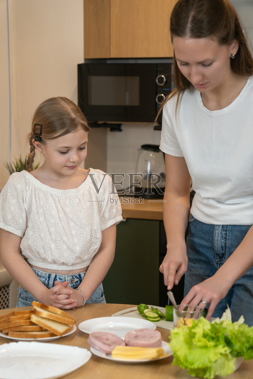 母亲在明亮的家庭厨房里准备三明治用的新鲜蔬菜，年幼的女儿在一旁观察，母女俩一起学习健康饮食和烹饪。竖幅照片照片摄影图片