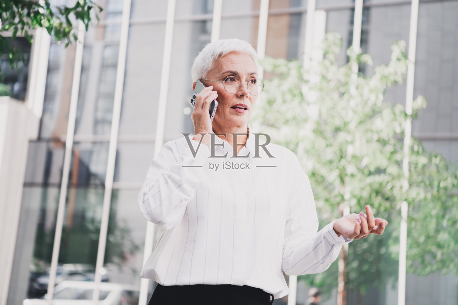 一位自信的老年女性穿着白衬衫，在阳光明媚的城市街道上，身旁是现代化的玻璃建筑，她正在打电话。照片摄影图片