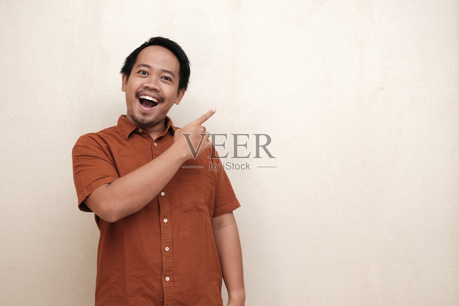 一位东南亚男子穿着棕色衬衫，面带微笑，热情地指向一旁，暗示有放置文字或宣传的空间。照片摄影图片