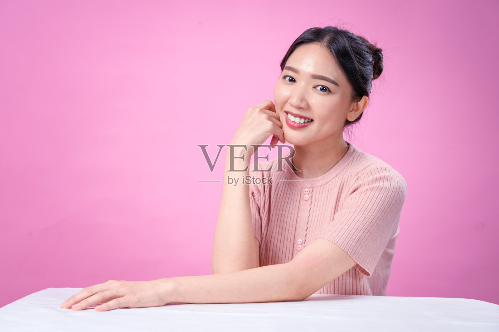 一位年轻的亚洲女性，梳着整洁的发髻，留着深色头发，身穿罗纹粉色短袖上衣，面带灿烂的笑容，手托着下巴，摆出愉快的姿势，背景是纯粉色。照片摄影图片