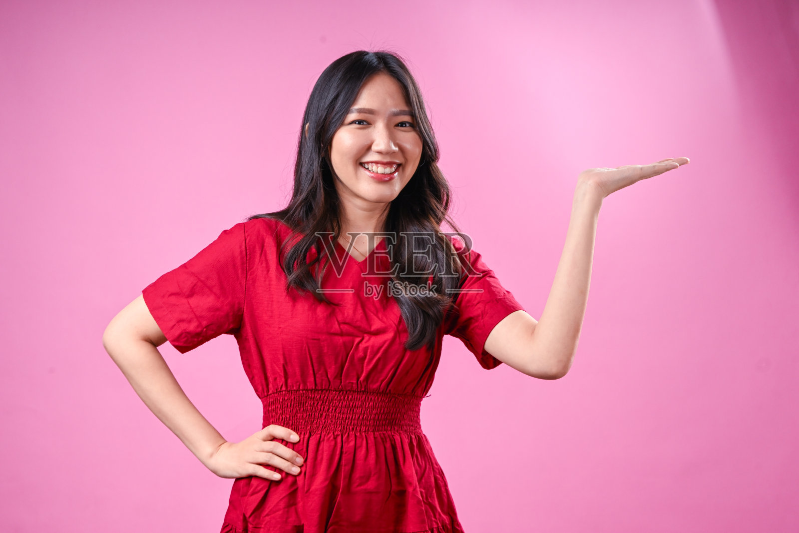 一位留着黑色波浪长发的年轻亚洲女性身穿红色连衣裙，在粉色背景前张开双手，灿烂地笑着，展现出兴奋和积极的情绪照片摄影图片