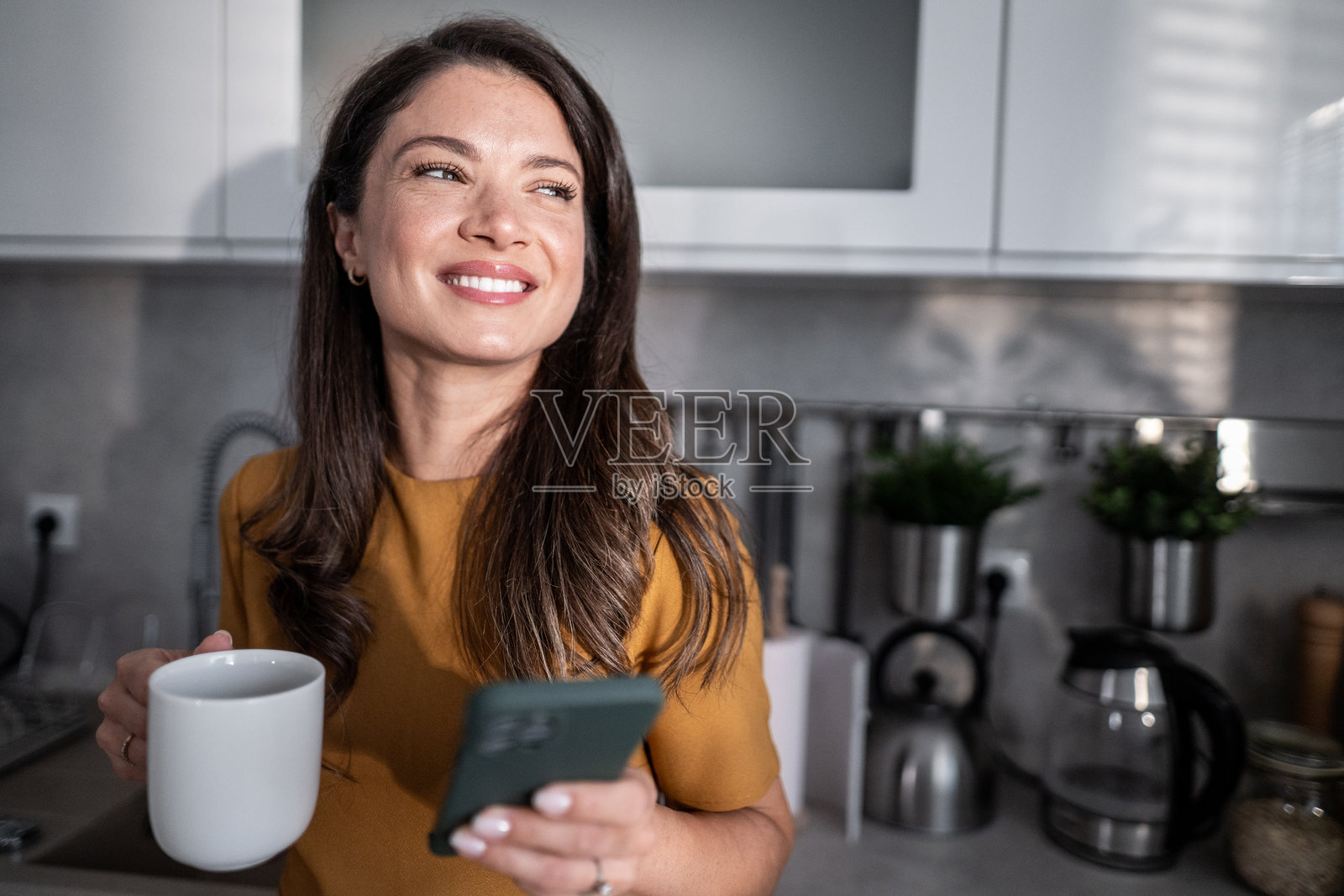 快乐的女人在厨房喝咖啡，同时使用智能手机照片摄影图片