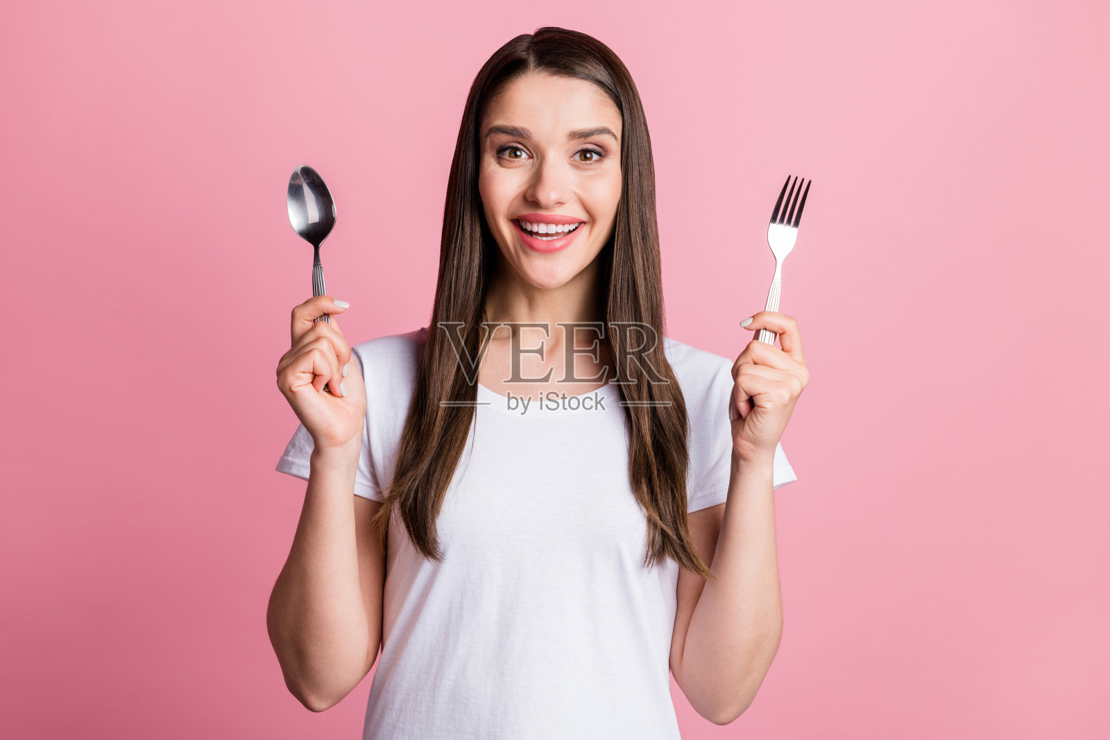 年轻女子微笑肖像，手拿勺子和叉子，等待午餐，背景为孤立的粉彩色。照片摄影图片