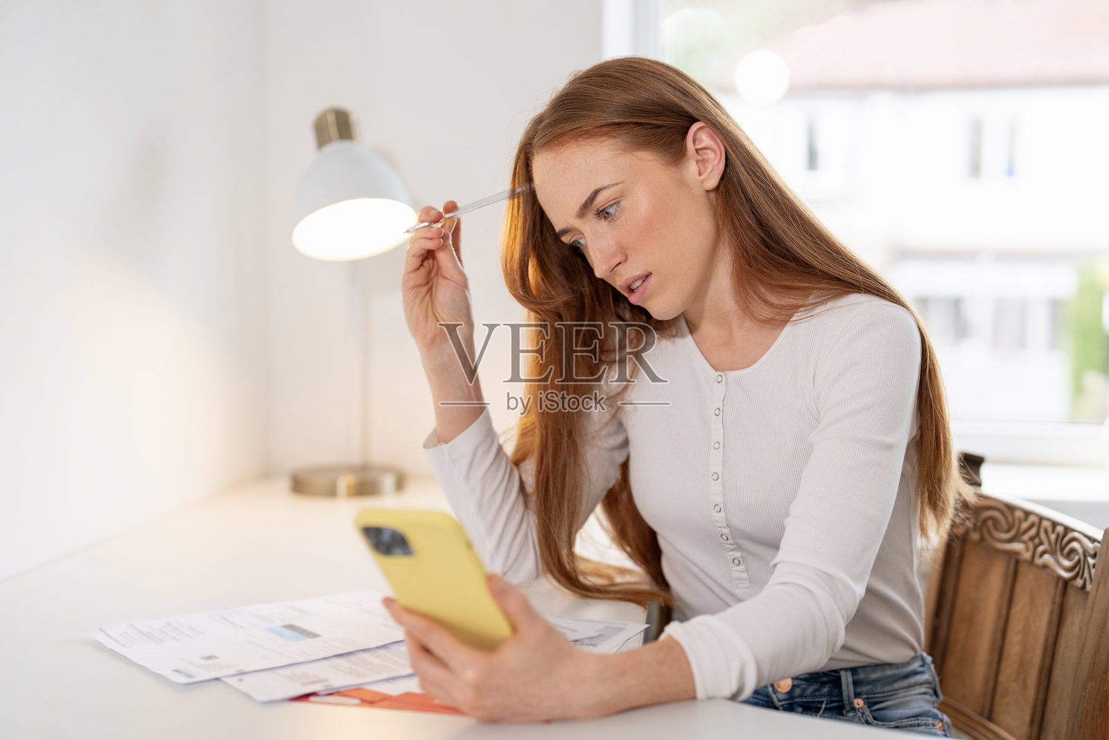 女子在家办公桌前，一边处理文件一边专注于智能手机照片摄影图片